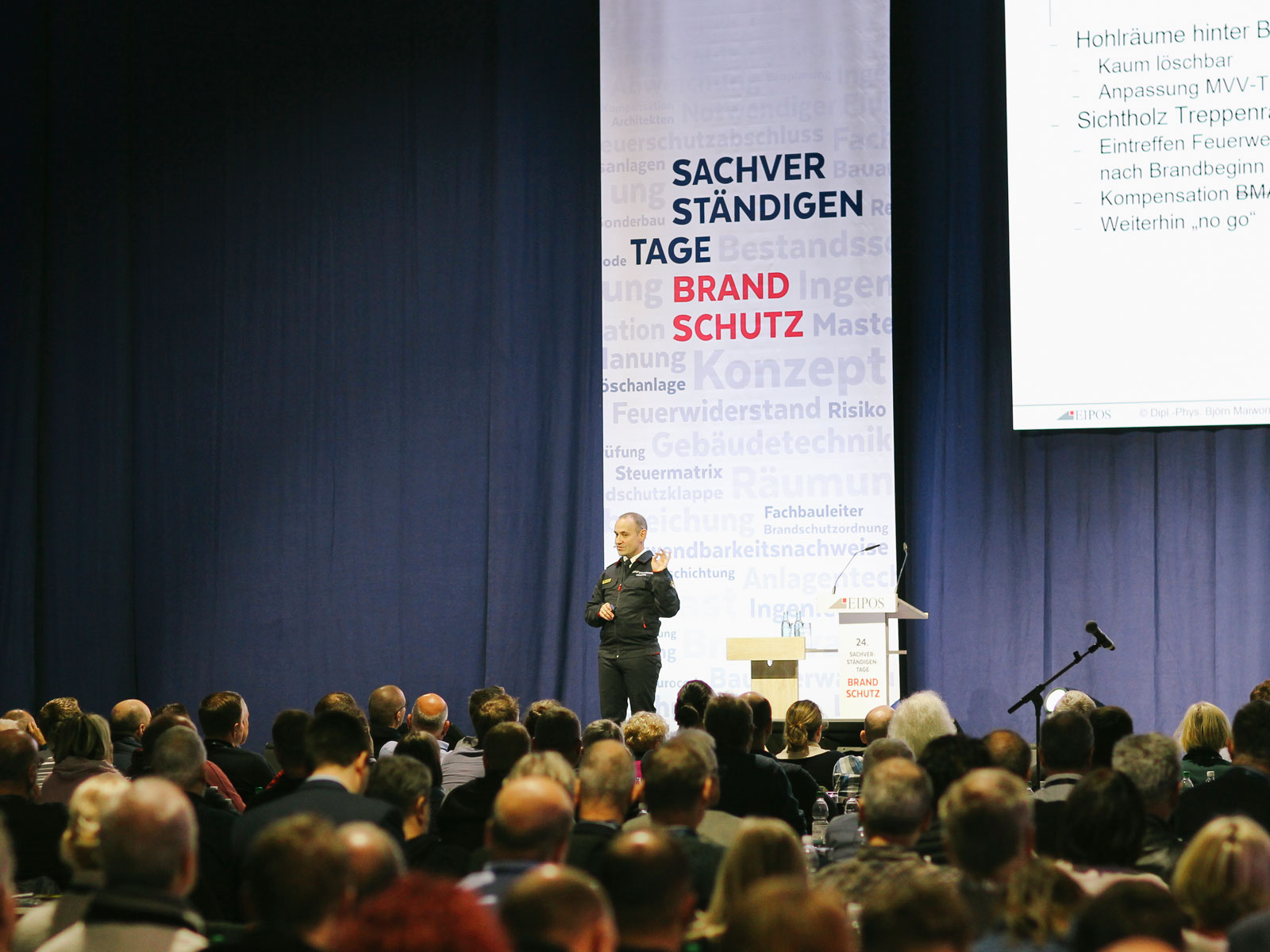 Foto eines Sprechers auf der Bühne der EIPOS Sachverständigentage Brandschutz. Das Publikum sitzt mit dem Rücken zur Kamera.