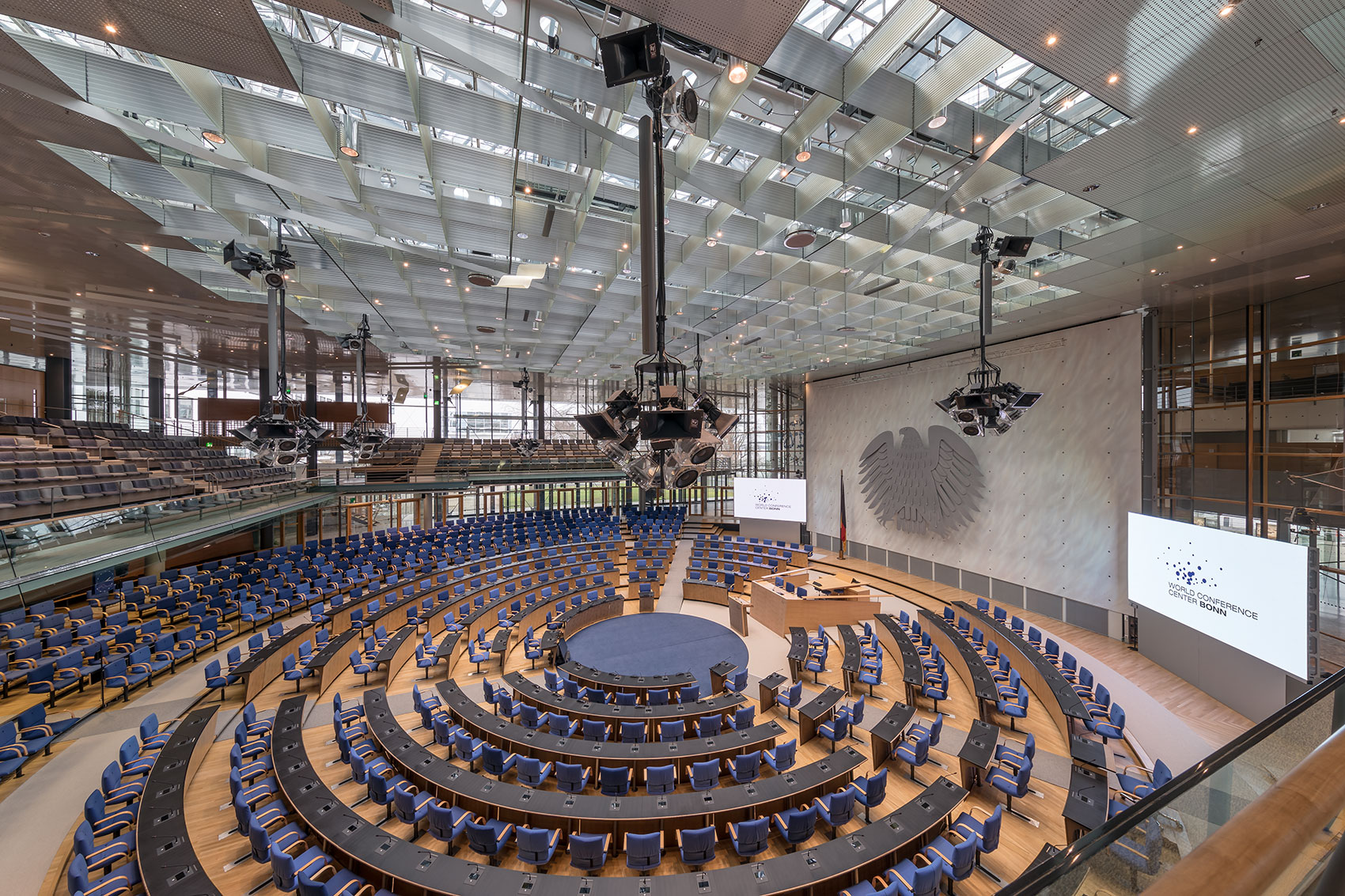 Konferenzsaal des World Conference Center Bonn: Viele in einem Kreis angeordnete Tisch- und Stuhlreihen. Licht- und Tontechniksäulen hängen von der Decke