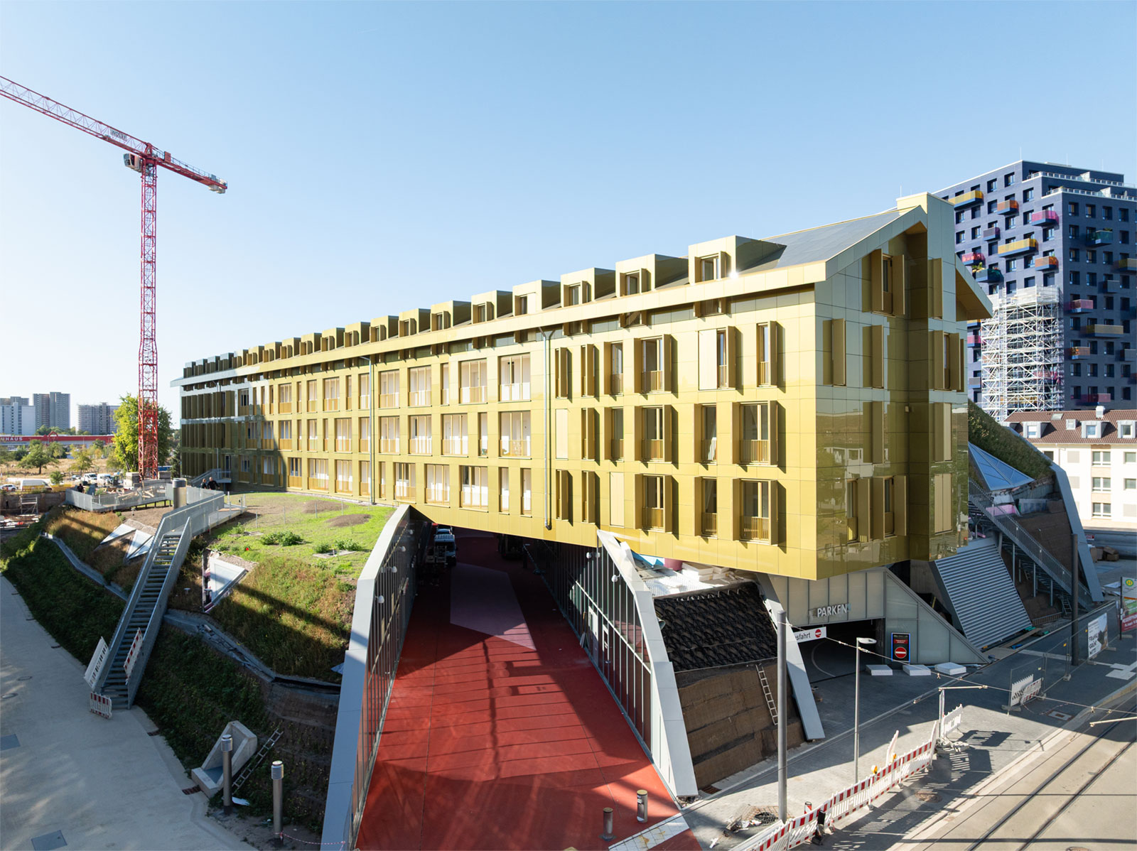 Neubau Grüne Mitte in Mannheim nach der Fertigstellung. Gelbes Haus auf einem künstlich angelegten Hügel. Unter dem Haus führt ein Fußgängerweg in das Nahversorgungszentrum.