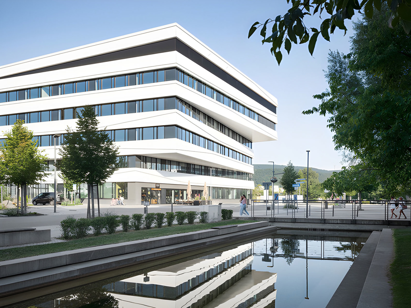 Ein fünfstöckiges Geschäftshaus mit weißer Fassade und langen Fensterstreifen über die ganze Breite spiegelt sich in einem Wasserbecken