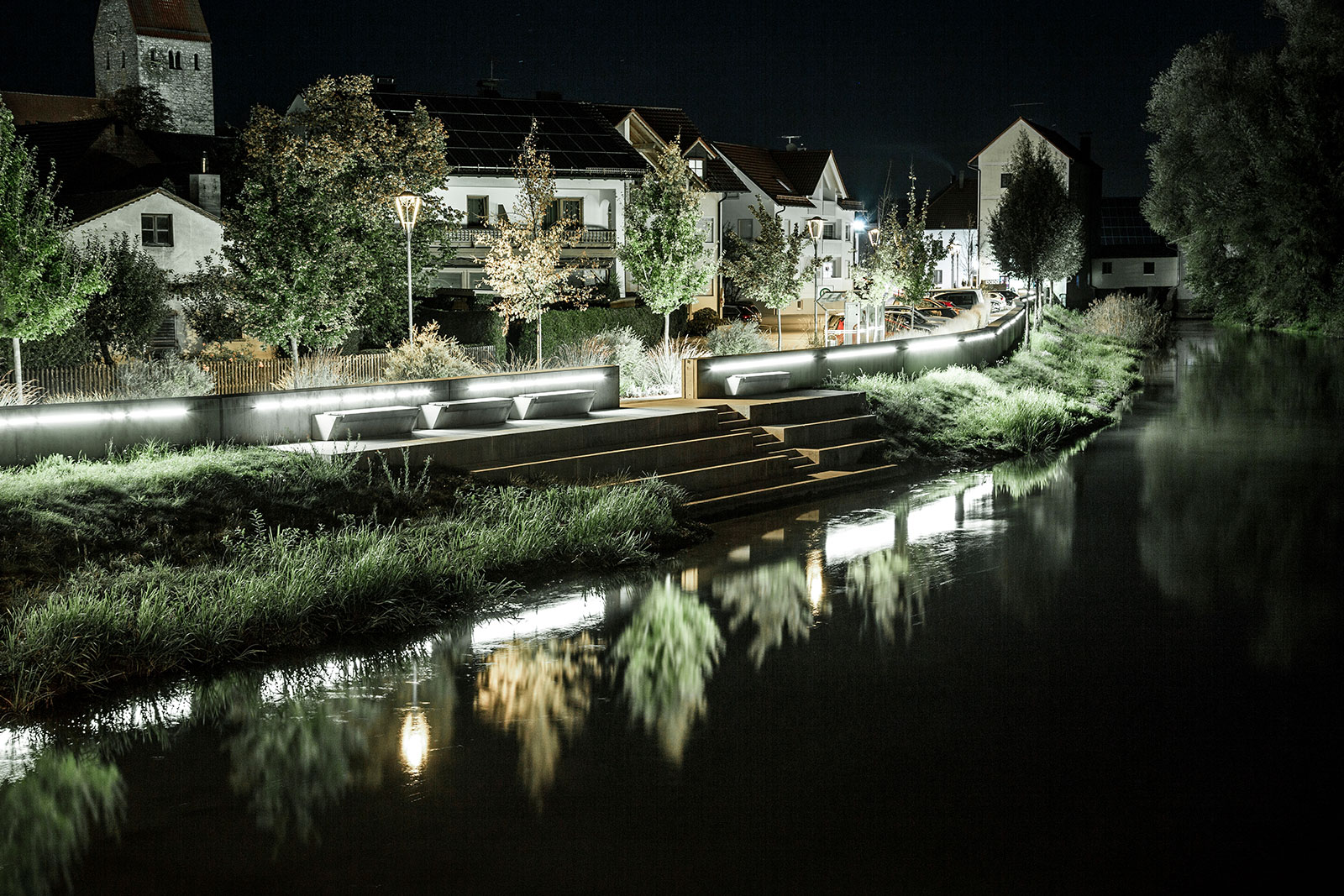 Das Profilbild von INKA Freiraum Design aus Ingolstadt zeigt ein beleuchtetes Wohnhaus bei nacht, das am Wasser liegt. Es spiegelt sich im Wasser.