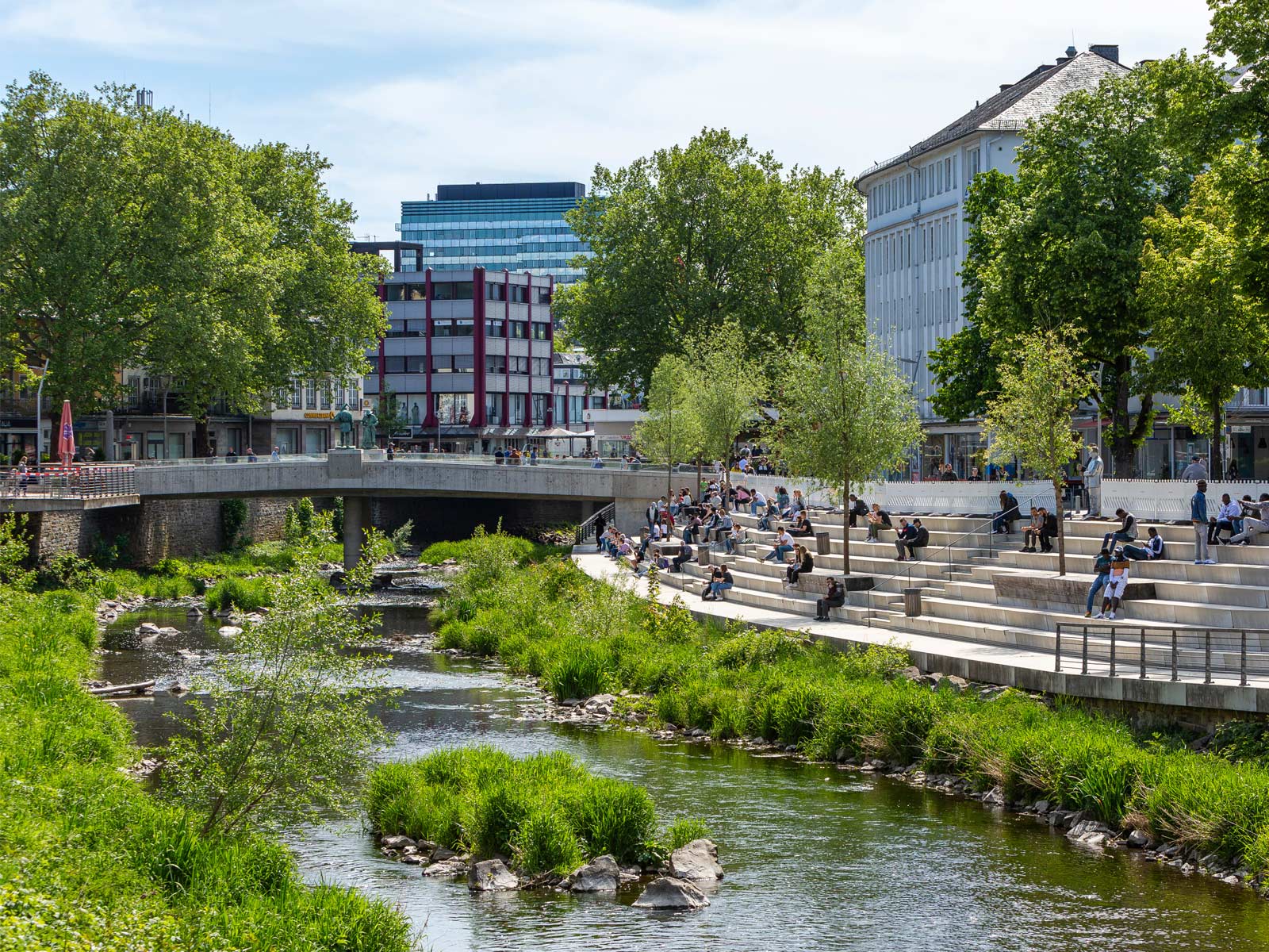 Das Beitragsbild zur neuen Förderung der KfW symbolisiert den Natürlichen Klimaschutz: Ein Fluss mit üppig grünen ufern verläuft durch eine Innenstadt. Menschen sitzen auf einer Treppe unter Schattenspendenden Bäumen.