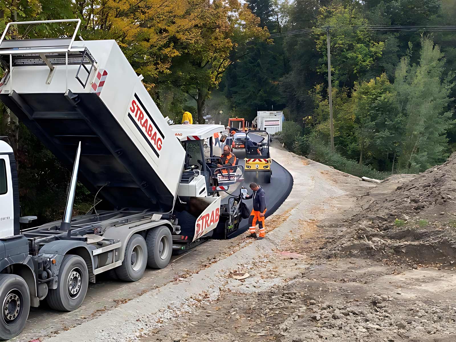 Bauarbeiter asphaltieren einen neuen Weg mit Biotumen. Baumaschinen tragen die Aufschrift STRABAG.