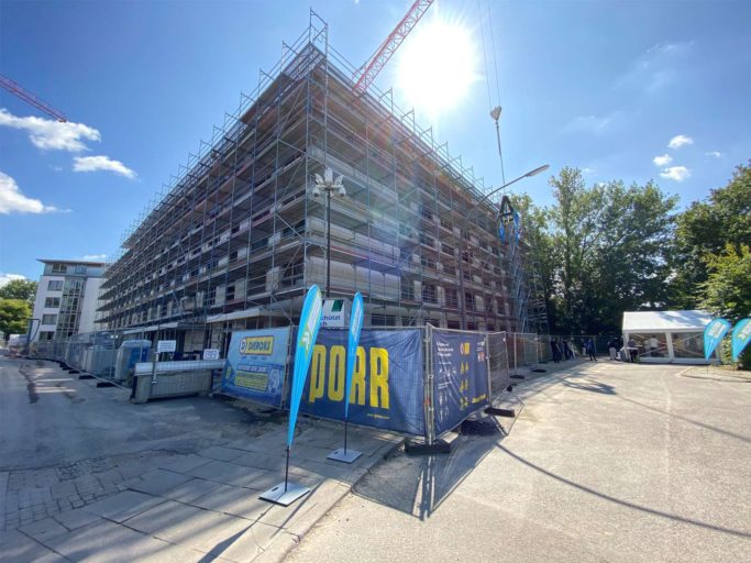 Der Rohbau der Pflegeeinrichtung PFLEGEN & WOHNEN WILHELMSBURG in Hamburg ist in Aufsicht gegen das Sonnenlicht fotografiert. Vor dem quadratischen Gebäude, das noch in Gerüste gehüllt ist, steht ein Bauzaun mit dem PORR-Logo.