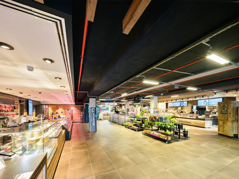 Supermarkt mit Frischetheken, Blumenregal und Bäckerei unter dunkler Deckenverkleidung