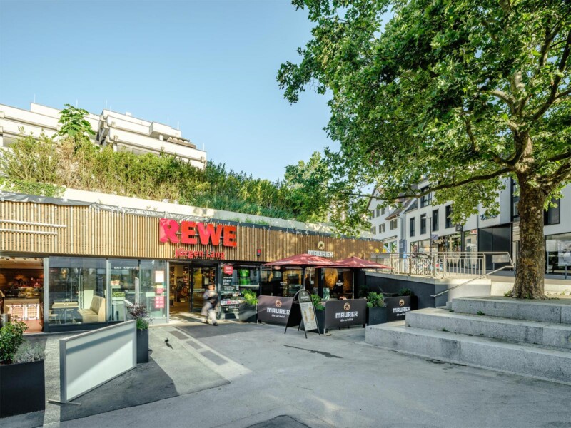 Rewe am Alten Postplatz in Waiblingen: Marktzugang mit Holzlamellenfassade und angrenzendem Außenbereich unter Bäumen