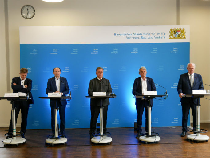 Fünf Männer stehen beim runden Tische der Baukonjunktur in Bayern an Rednerpulten vor einem Hintergrundbanner, das das Logo des Bayrischen Staatsministeriums für Wohnen, Bau und Verkehr zeigt