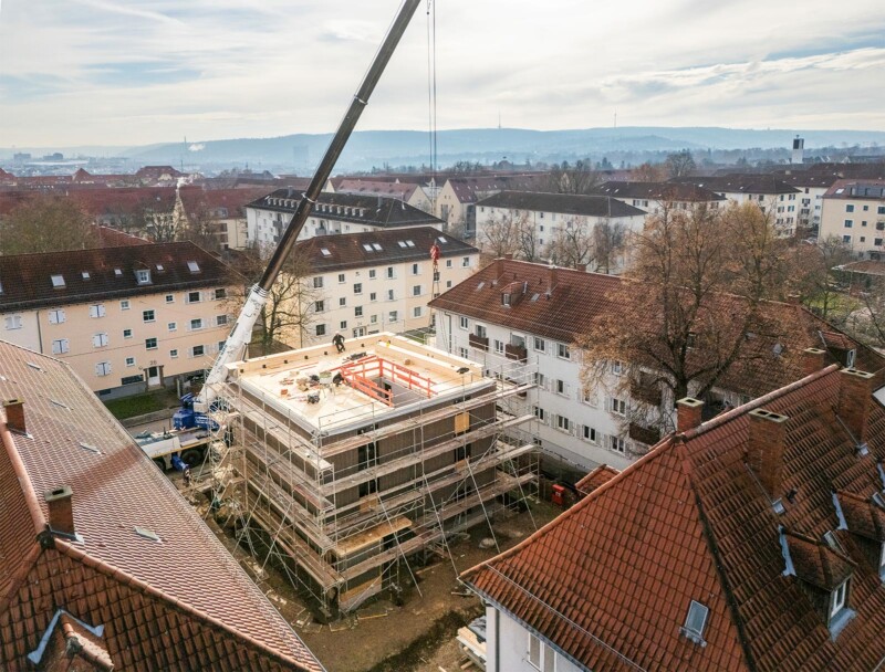 Baustelle eines Holzbaus mit Baukran im Innenhof einer Wohnsiedlung mit roten Ziegeldächern