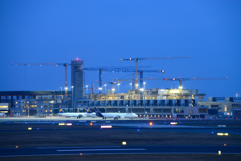 Die baustelle des Terminal 3 bei der Dämmerung. Viele Baukräne zieren den tiefblauen Nachthimmel. Im Vordergrund stehen Flugzeuge.