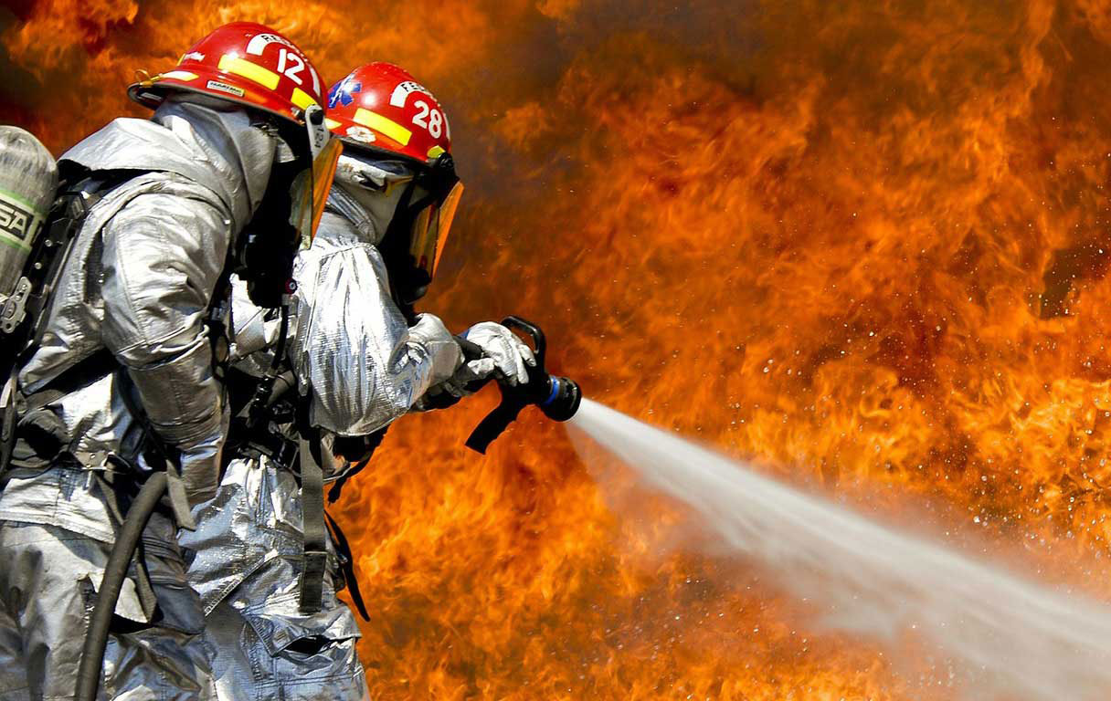 Auf dem Profilbild von TOP Brandschutz aus Stuttgart sind zwei Menschen in Hitzeschutzanzügen zu sehen, die ein Flammenmeer mit einem Wasserstrahl bekämpfen.
